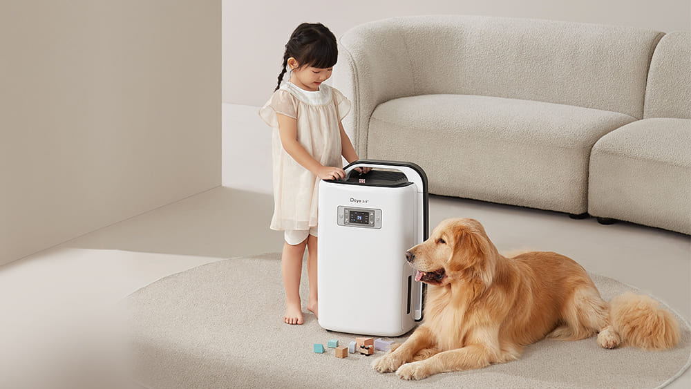 A young girl interacts with a portable dehumidifier in a living room while a golden retriever watches nearby.