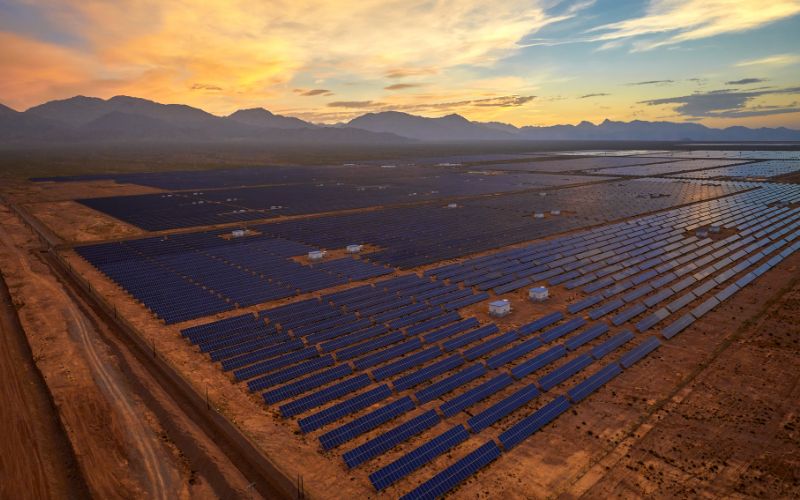 Aerial view of a vast solar farm with rows of photovoltaic panels at sunset, set against a backdrop of distant mountains and a cloudy sky.