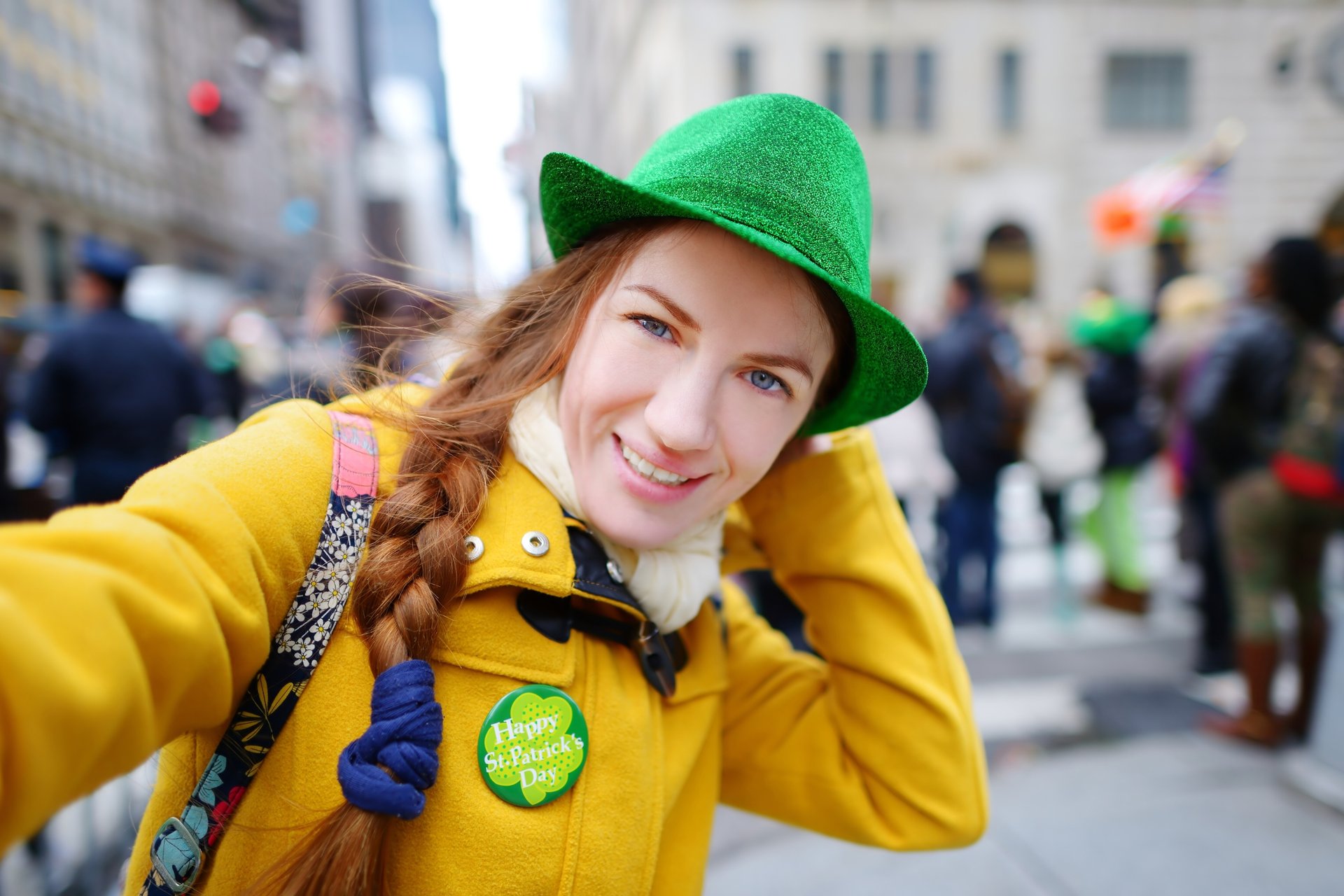 A young woman with a side braid and a bright smile is wearing a green leprechaun hat and a yellow coat,  taking a selfie.