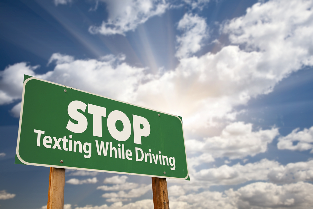 "Stop Texting While Driving" - A vivid green road sign against a backdrop of a dramatic sky, clouds, and sun