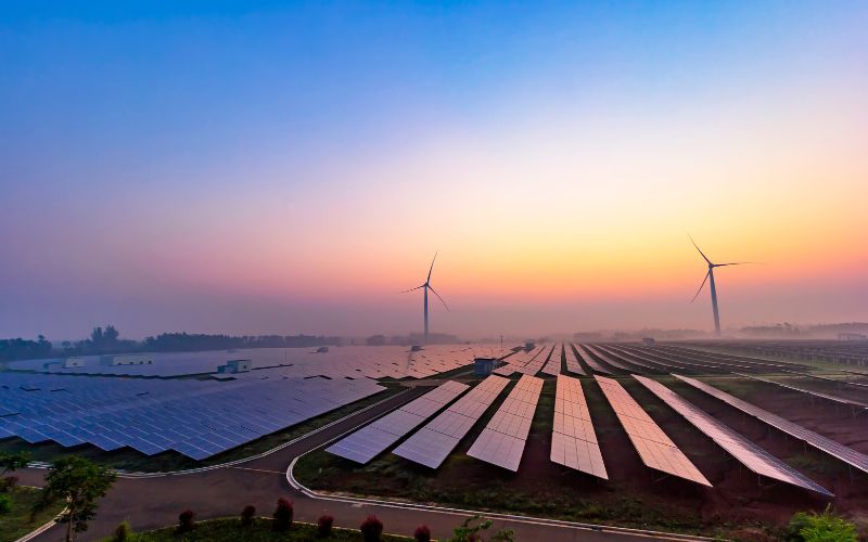 A sunrise over a renewable energy farm with rows of solar panels and wind turbines under a colorful sky.