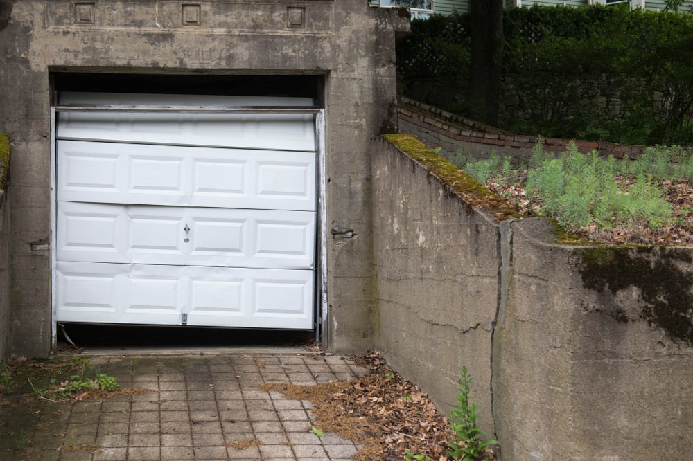 A single garage door, askew and dented, set against a backdrop of a property that looks neglected, with surfaces covered in old leaves and debris.