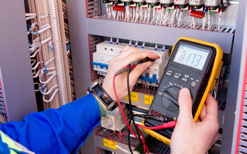 An electrician using a multimeter to measure voltage in a complex industrial electrical panel.